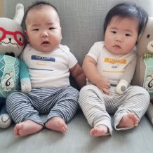 Twin asian 6-month-old babies wearing custom Hello My Name is simpelwear bodysuits, with stuffed animals.